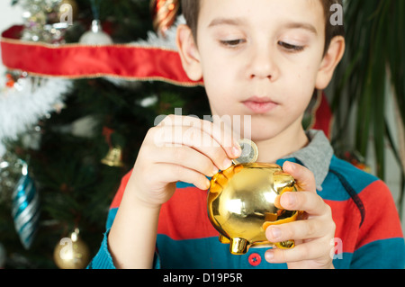 Kleiner Junge legt eine Münze in Bargeld Schwein vor dem Christmass Baum. Stockfoto