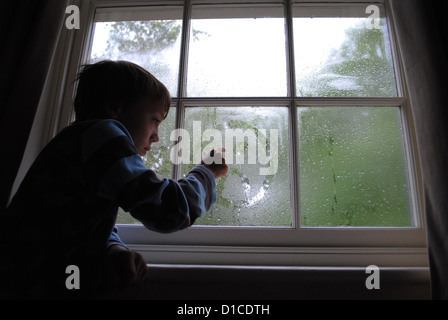 8 Jahre alter Junge zeichnen ein trauriges Gesicht in das Kondenswasser am Fenster. Stockfoto