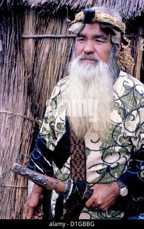 Ein bärtiger Ainu-Mann im traditionellen Kleid und das tragen einer Holzfaserkrone posiert 1962 mit einem zeremoniellen Schwert außerhalb seines Schilfhauses in Hokkaido, Japan. Stockfoto