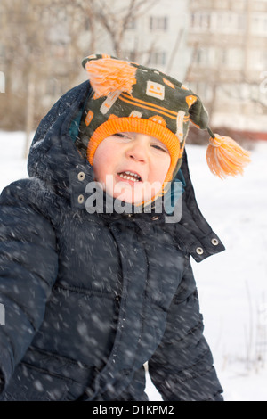 Niedlichen kleinen Jungen mit einer warme Wollmütze mit Kordeln gefangen in einem Schneegestöber hinuntertreiben vorbei sein Gesicht als Schneeflocken an einem eiskalten Wintertag. Stockfoto