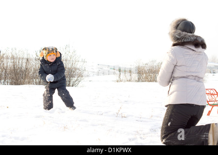 Kleiner Junge stehend in einem verschneiten Feld werfen Schneebälle auf seine Mutter, die sich mit dem Rücken zur Kamera kniet Stockfoto