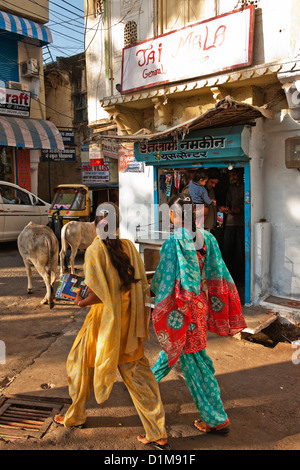 Zwei indische Mädchen in leuchtenden Saris zu Fuß auf einer typischen indischen Straße mit Ladengeschäft und zwei Kühe an einem heißen sonnigen Tag in Udaipur Stockfoto
