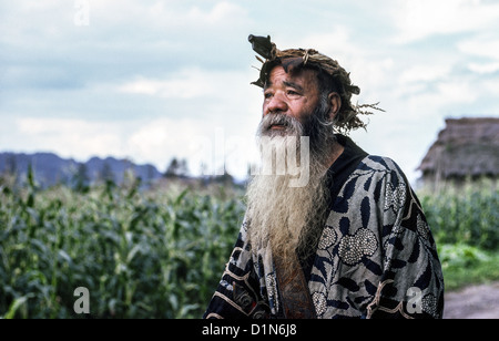 Ein älterer Ainu-Mann mit langem Bart und einer Krone aus Holzfasern posiert im traditionellen Kleid außerhalb seines ländlichen Hauses in Hokkaido, Japan.i. Stockfoto