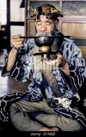 Ein älterer bärtiger Ainu-Mann verwendet einen Holzheber, um seinen Schnurrbart aus einem Trinkbecher in Hokkaido, Japan, Ostasien zu halten. 1962 fotografiert. Stockfoto