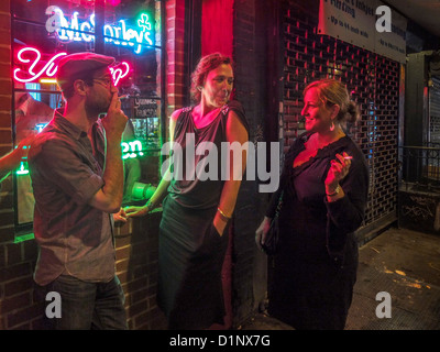 Ein Mann und zwei Frauen rauchen auf dem Bürgersteig vor einem Restaurant in der Nacht in New Yorks Lower East Side. Stockfoto