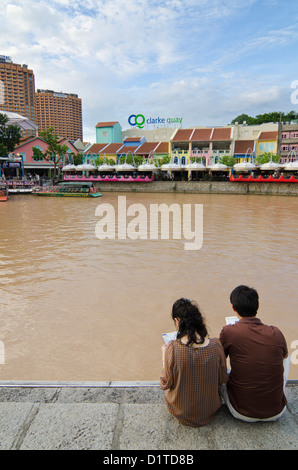 Ein paar sitzen am Ufer Singapur-Flusses, die mit Blick auf die bunt bemalten Unterhaltungsviertel von Clarke Quay Stockfoto