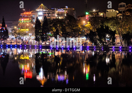 HANOI, Vietnam – die Lichter der Altstadt von Hanoi spiegeln sich nachts auf dem Hoan Kiem See (Ho Hoan Kiem). Die Mauer des Sees an diesem Ende ist von Scheinwerfern umgeben, die ihre Farbe ändern. Dahinter befinden sich die Lichter von Touristenbars und Restaurants mit Blick auf den See. Stockfoto