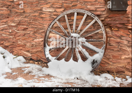 Eine alte, antike Wagenrad mit Schnee bedeckt ruht einer Ranch Anlage Wand. Stockfoto