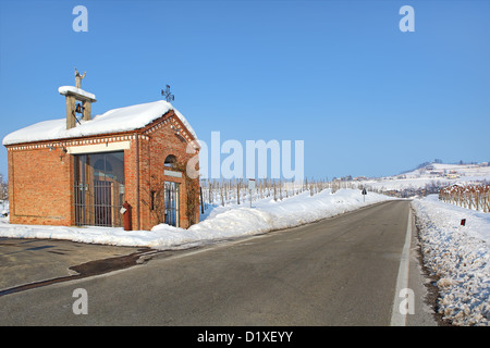 Kleine Rote Kapelle am Straßenrand zwischen Weinbergen, die unter dem weißen Schnee unter klaren, blauen Winterhimmel im Piemont, Norditalien. Stockfoto