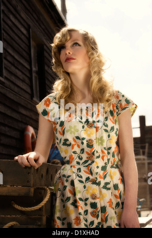 Junge blonde Frau in Vintage print Kleid gelehnt Stapel von hölzernen Auster Tabletts mit Seil Griffe im sonnigen Kai Lage Stockfoto