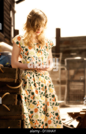 Junge blonde Frau in Vintage print Kleid gelehnt Stapel von hölzernen Auster Tabletts mit Seil Griffe im sonnigen Kai Lage Stockfoto