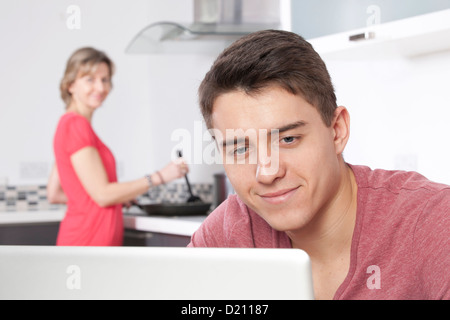 Junger Mann mit einem Laptop, ist eine Frau im Hintergrund kochen. Stockfoto