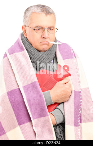 Krank Senior mit Thermometer im Mund, bedeckt mit Decke hält eine Wärmflasche isoliert auf weißem Hintergrund Stockfoto