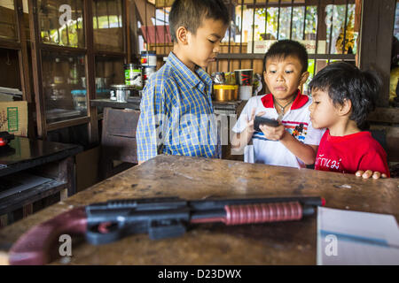 13. Januar 2013 spielen - Bangkok, Thailand - jungen mit einem Smartphone, während ihre Spielzeugpistole im Stadtteil Bang Luang Bangkok vor ihnen sitzt. Stadtteil Bang Luang Linien Khlong (Kanal) Bang Luang in der Thonburi-Sektion von Bangkok auf der westlichen Seite des Chao Phraya Flusses. Es wurde Ende des 18. Jahrhunderts von König Taksin dem großen gegründet, nachdem die Birmanen die siamesische Hauptstadt Ayutthaya entlassen. Die Nachbarschaft, wie die meisten von Thonburi, ist relativ unerschlossen und noch Kreuz und quer durchzogen von den Kanälen, die einmal Bangkok berühmt gemacht. Es ist jetzt ein beliebter Tagesausflug vom Zentrum von Bangkok ein Stockfoto