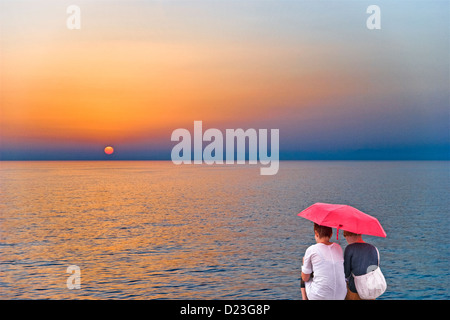 Zwei Damen sitzen unter einem Sonnenschirm mit einem entspannenden Chat im Regen, Sonne einstellen / über dem Meer aufgeht. Manipulierte Bild Stockfoto