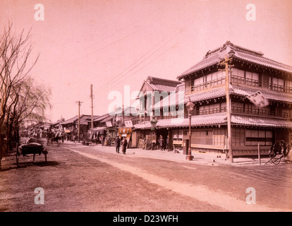 Straßenszene in der Stadt mit gewerblichen und/oder Wohn Gebäude entlang einer Seite und Straßenlaternen und Strommasten; ein paar, im Zentrum, mitten auf der Straße. Yokohama, Japan, um 1890 Stockfoto