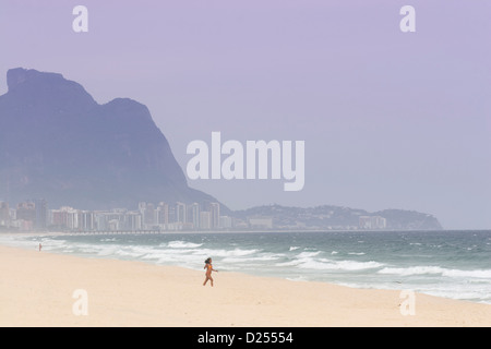 Pontal, Barra da Tijuca, Rio. Eine junge Frau in einem Bikini, die mit dem Berg Pedra da Gavea und der Skyline der Stadt in der Ferne in die Wellen läuft Stockfoto