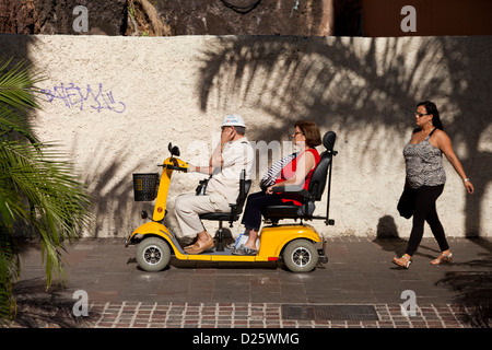 Paar auf einem Doppelsitzer-Roller für behinderte Menschen auf dem Bürgersteig in Los Cristianos, Teneriffa, Kanarische Inseln, Spanien. Stockfoto