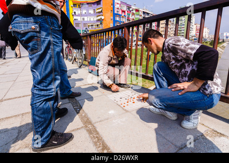 Junge Männer spielen Dame auf dem Boden an einer Brücke über den Fluss Lana mit bunten Gebäuden im Hintergrund. Tirana, Albanien Stockfoto