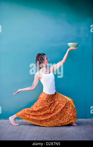 Einzigartige Frau in eine tanzende Yogastellung bietet sich eine schöne Keramikschale vor dem Hintergrund einer blauen Wand. Stockfoto