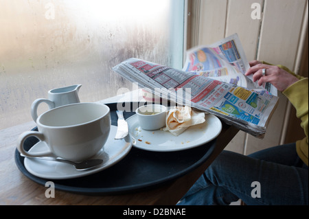 Frau liest Zeitung am Café Fenster mit leeren Becher und Teller, England, UK Stockfoto