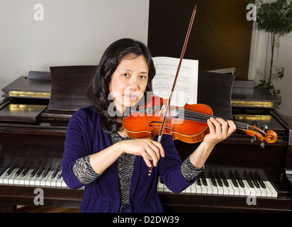 Reife Asiatin, Geige zu spielen, mit alten dunklen hölzernen Klavier im Hintergrund Stockfoto