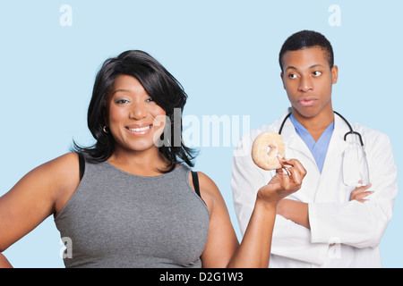 Übergewicht Mischlinge Frau mit Donut von Arzt über blauem Hintergrund Stockfoto