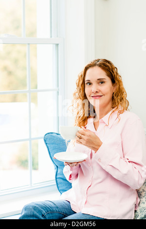 Lächelnde kaukasischen Frau entspannend auf Couch von Fenster halten Tasse Kaffee Stockfoto