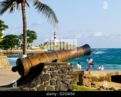 Menschen entspannen von einer alten Canon mit Ferol Leuchtturm im Hintergrund am Strand von Salvador, Brasilien Stockfoto