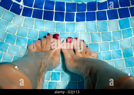 Frauenfüße mit rot bemalten Zehennägeln auf blauen Schwimmbadfliesen unter Wasser Stockfoto