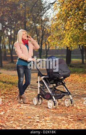 Junge Mutter sprechen auf einem Handy bei einem Spaziergang im Park mit ihrem Baby im Kinderwagen Stockfoto