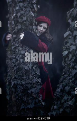 eine Frau in einem roten Mantel ist einen alten Baum mit Efeu umarmt. Stockfoto