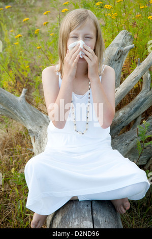 Junges Mädchen sitzen auf einem Baumstamm im Sommer umgeben von Unkraut und bläst ihre Nase wegen einer Allergie Stockfoto