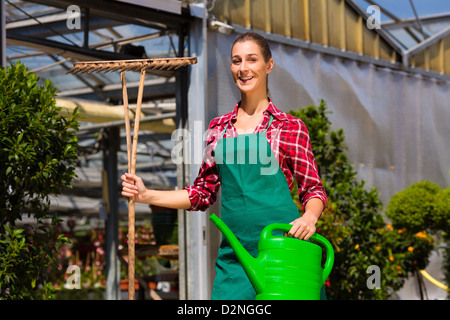 Weibliche gewerbliche Gärtner vor grünen Haus mit Topf gießen und Rechen Stockfoto