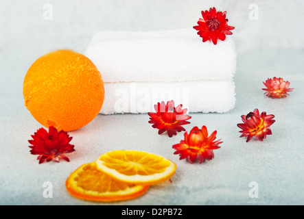 Zwei weiße Frottierwäsche, dekoriert mit orangen Blüten, getrocknete orange Kugel neben der Badewanne, im Vordergrund orange Segmente Stockfoto