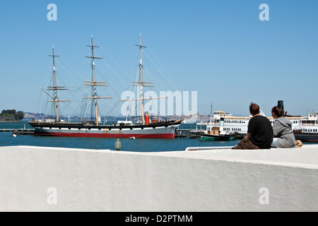 Junge Paare auf einer Hafenmauer in Richtung historische Schiffe am Hyde Street Pier in der Nähe von Ghirardelli Square in San Francisco, Kalifornien suchen Sitzen Stockfoto