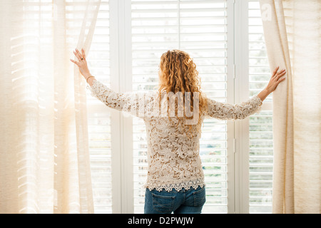 Frau, die aus großen hellen Fenster mit Vorhängen und Jalousien Stockfoto