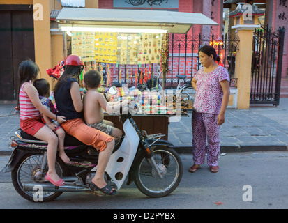 Kinder vorbeischauen auf ihr Motorrad anzusehen Schmuckstücke verkauft werden, von einer Frau mit einem Push Cart, Hoi an, Vietnam, Indochina Stockfoto