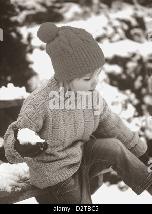Foto im Vintage-Stil eines jungen Mädchens, das Pullover und Hut im Vintage-Stil trägt und im Winter einen Schneeball in Cornwall, England, hält Stockfoto
