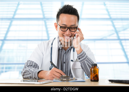Southeast Asian junge Arzt schreiben, während auf dem Handy anrufen sitzt vor einen Schreibtisch im Krankenhaus. Stockfoto