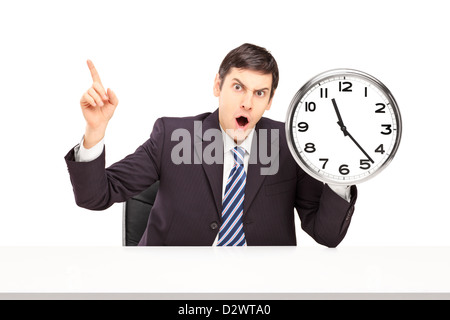 Wütend Geschäftsmann sitzen und halten eine Wanduhr isoliert auf weißem Hintergrund Stockfoto