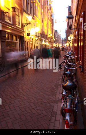 Eine belebte Straße von Amsterdam in der Abenddämmerung mit ikonischen Fahrräder geparkt entlang der Seite, während Menschen Geschäfte, Cafés und Bars besuchen. Stockfoto