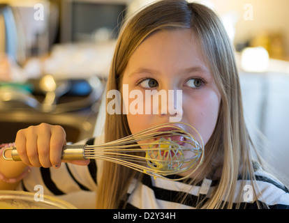 Junge Mädchen leckt den Löffel Schneebesen, während sie Kuchen in einer Küche mit Bokeh Stockfoto