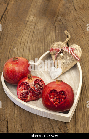 Zwei ganze und halbe Granatapfel auf einem Tablett mit einem Herz aus Holz, einen Granatapfel mit herzförmigen Ausschnitt Stockfoto