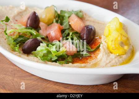 Ein Kurs von Hummus, serviert in einer weißen Schale auf einem Holztisch, garniert mit Olivenöl, Salat, Oliven, gewürfelte Tomaten Stockfoto