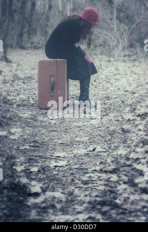 eine Frau mit einem schwarzen Mantel und eine rote Mütze sitzt auf einem alten Koffer im Wald Stockfoto