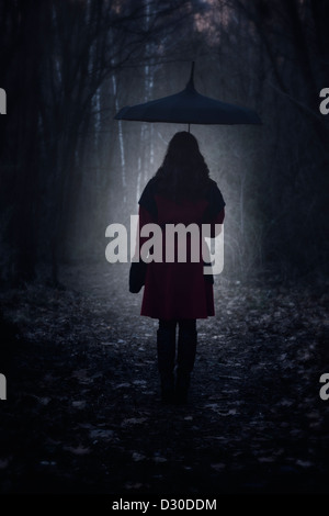 eine Frau mit einem roten Mantel und Regenschirm spaziert durch einen dunklen Wald Stockfoto