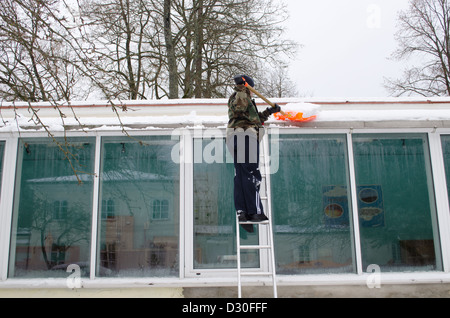 männlichen Mann in Tarnung auf Leiter sauber Schnee vom Dach des Hauses mit Kunststoff Schaufeln Werkzeug im Winter. Stockfoto