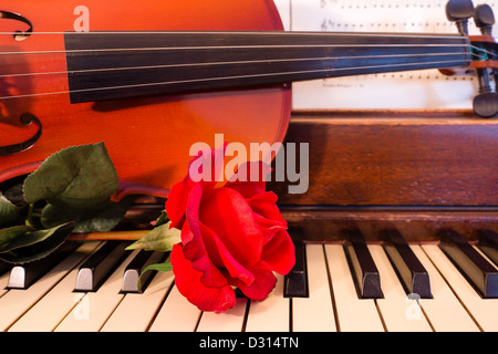 Eine einzelne rote Rose ruht auf einer Tastatur eines Klaviers zusammen mit einer Violine und Noten. Stockfoto