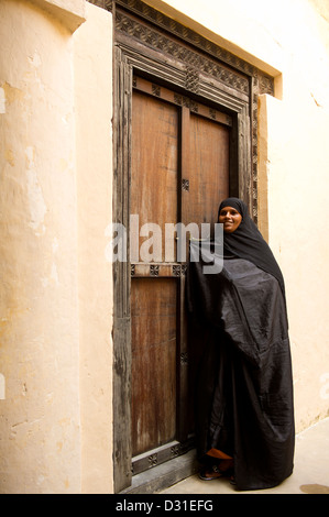 Swahili-Frau steht an einer traditionellen Zanzibar-Tür in der alten Festung, Lamu, Lamu Archipel, Kenia Stockfoto